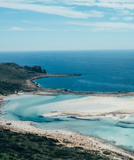Blick von oben auf eine weitläufige, helle Sandlagune mit flachem, türkisblauem Wasser, die an ein tiefblaues Meer grenzt.