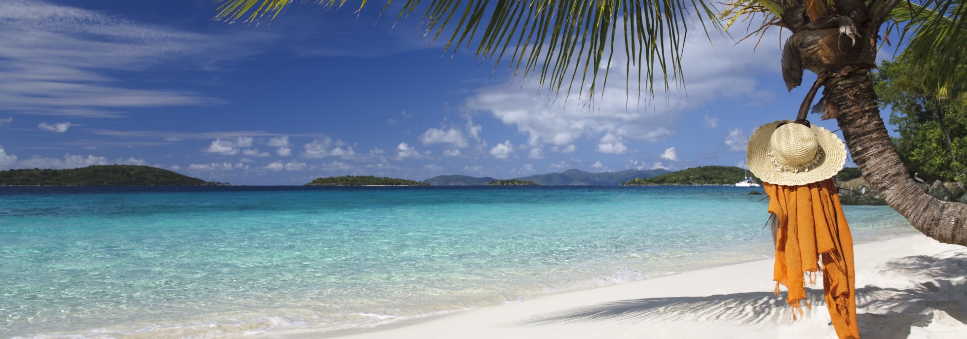 Ein malerischer, unberührter Sandstrand mit strahlend türkisblauem Meer und einem wolkenlosen, blauen Himmel. Eine Palme ragt von rechts ins Bild, an der ein Strohhut und ein orangefarbenes Handtuch hängen. Im Hintergrund erstrecken sich mehrere grüne Inseln am Horizont, was eine idyllische tropische Atmosphäre schafft.